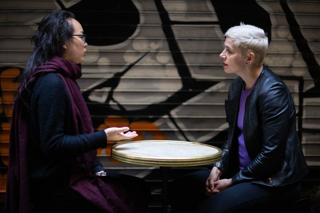 Tathra and a client at a table with graffiti in the background. A woman with dark hair, a scarf and glasses is speaking. Tathra is listening, wearing a black leather jacket and purple t-shirt. 
